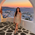 A woman wearing a shimmering champagne gold asymmetrical one-shoulder dress stands on a white stone walkway in front of a scenic Mediterranean coastal town at sunset, framed by a large white archway.